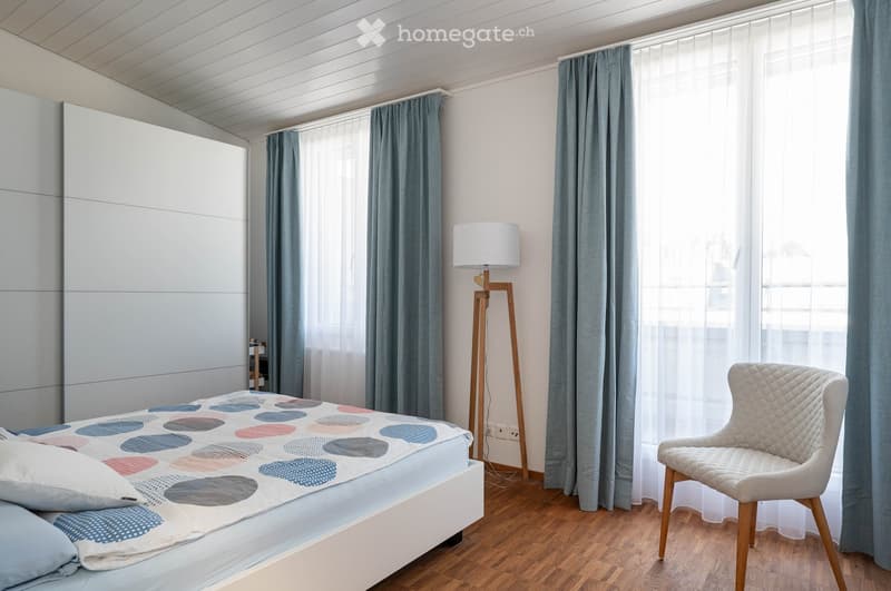 Modern bedroom with a bed, colorful quilt, white chair, floor lamp, and large windows with curtains.