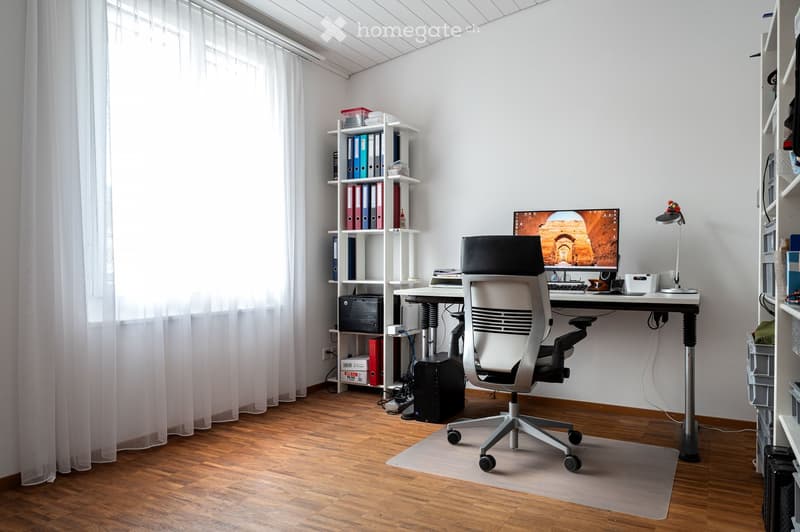 Modern office space with a wooden floor, large window with white curtains, ergonomic chair, desk with computer setup, shelves with binders and office supplies.