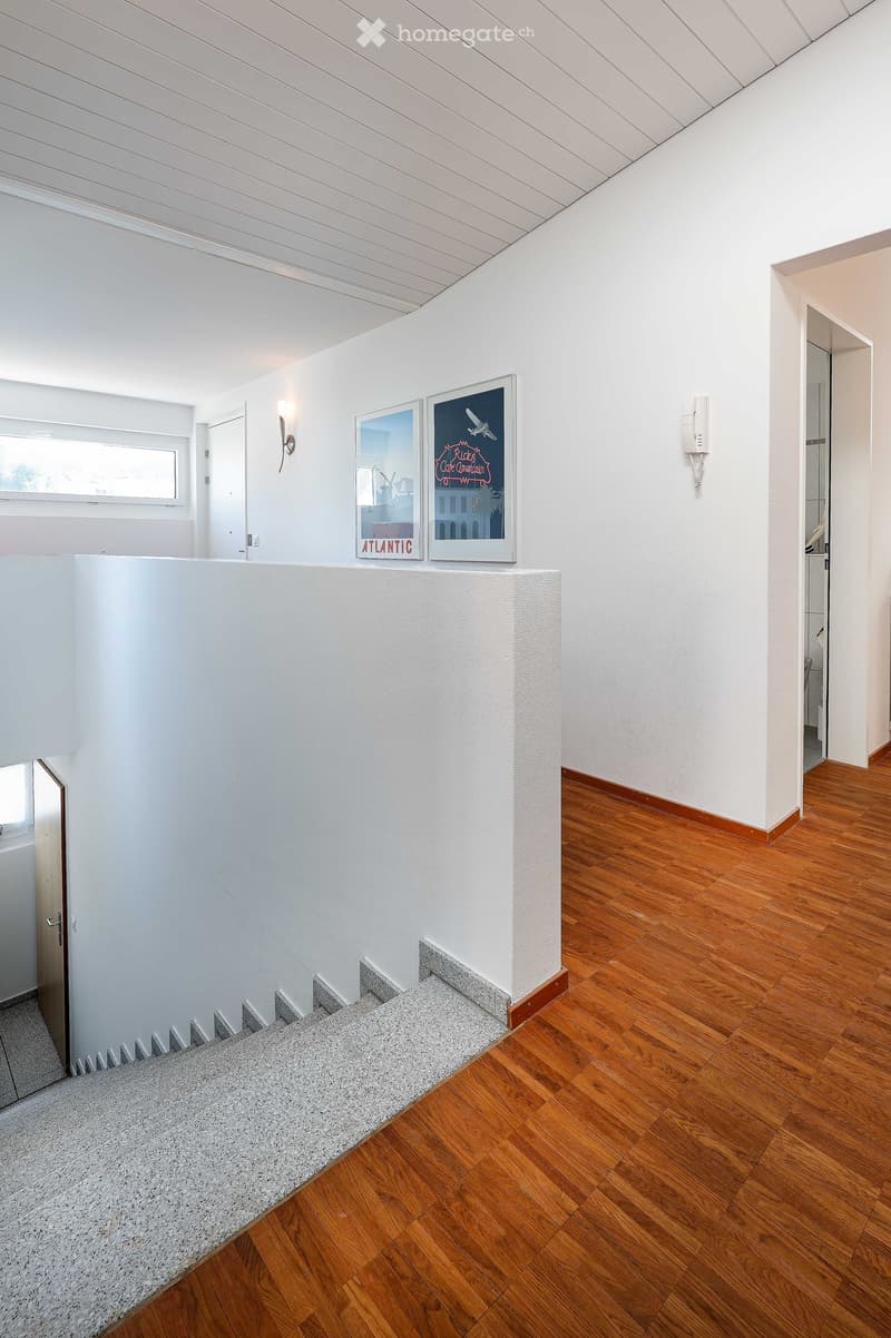 Modern hallway with wooden flooring, white walls, and a staircase leading upwards. Features include a wall-mounted light fixture, framed artwork, and a telephone on the wall.