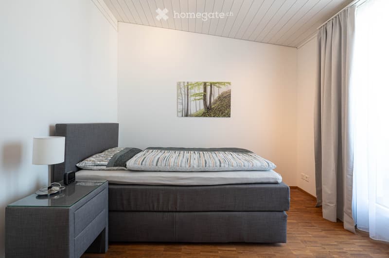 Modern bedroom with a grey bed, nightstand, lamp, and a picture frame on the wall. Wooden flooring and white walls.