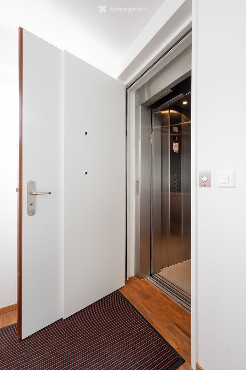 Hallway with wooden flooring, white walls, elevator access, and a closed door.