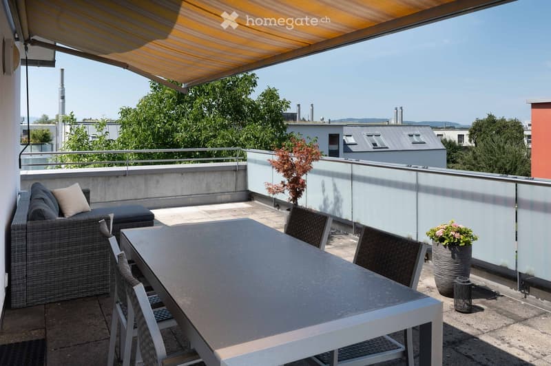 Balcony with a covered area, featuring a sofa, a dining table with chairs, and a flower pot. The balcony has a modern design with a glass railing and a view of greenery and buildings.