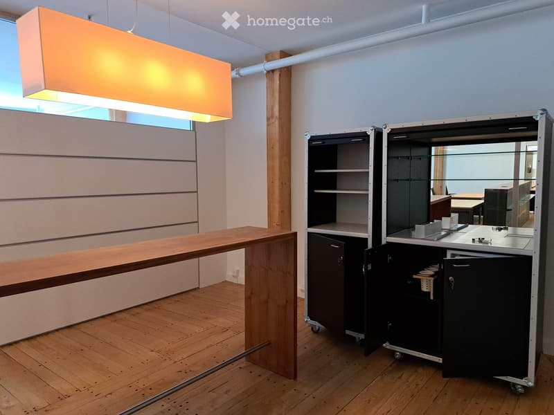 Modern, minimalist room with a long wooden counter, a black cabinet with glass doors, and a sleek light fixture hanging from the ceiling.