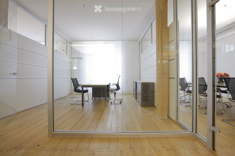 Modern office space with wooden flooring, glass partitions, and contemporary furniture including desks, chairs, and a small table with a flower vase.