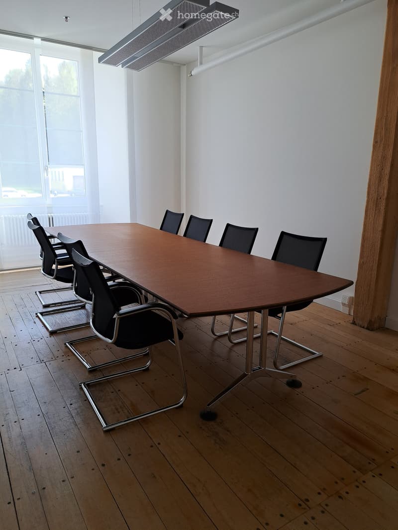 Conference room with a large wooden table and multiple black chairs arranged around it. The room has wooden flooring and large windows allowing natural light to enter.