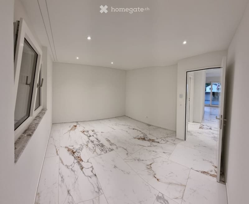 Empty room with white walls, marble flooring, and recessed lighting. Features a large window and an open doorway leading to another space.