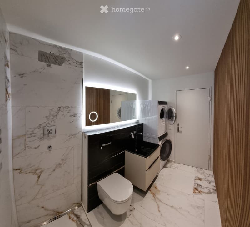 Modern bathroom with marble tiles, a toilet, sink, mirror, shower, and stacked washer/dryer unit.