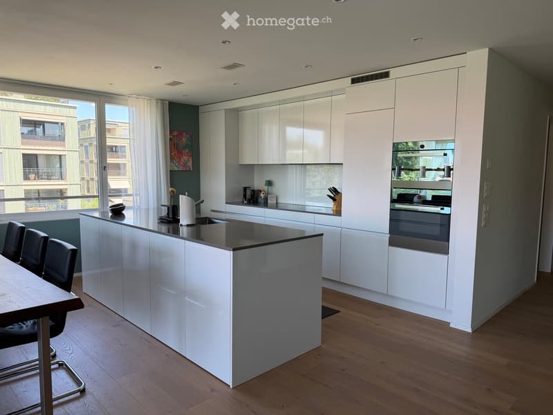 Modern kitchen with white cabinets, island, and stainless steel appliances. Features a dining area with a table and chairs, and a large window with a view of the building outside.