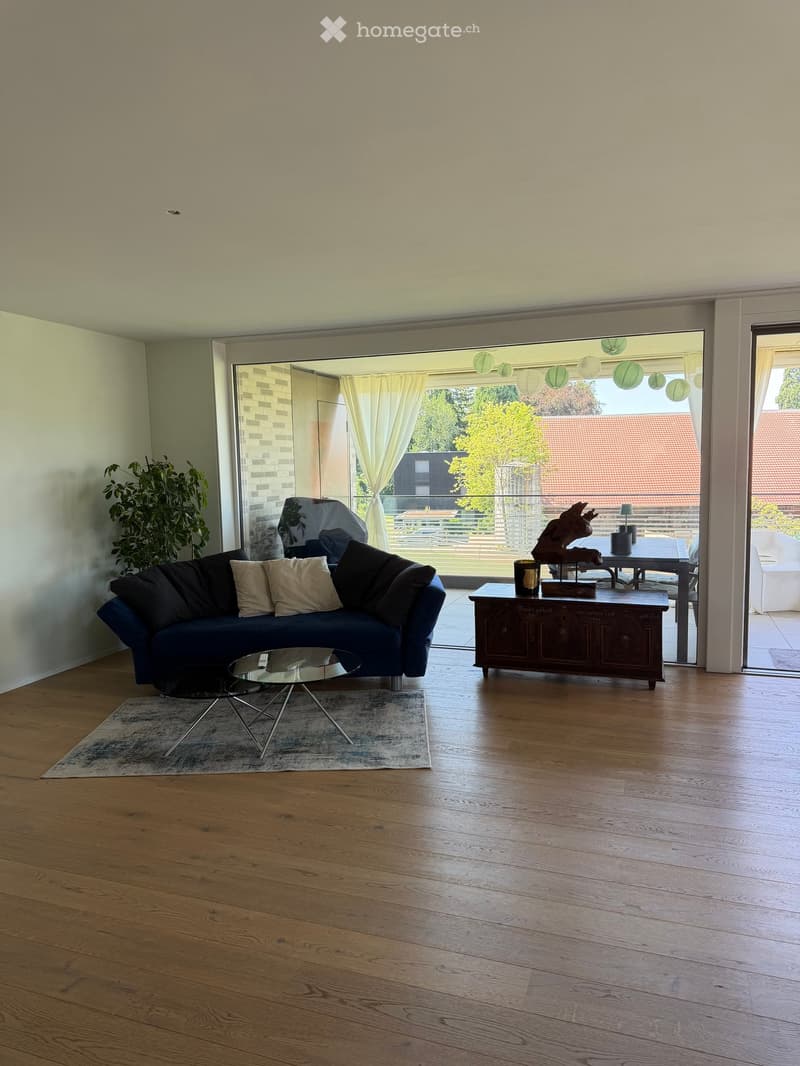 Modern living room with wooden flooring, large windows, and a balcony view. Features a blue sofa, glass coffee table, side table, and decorative items.