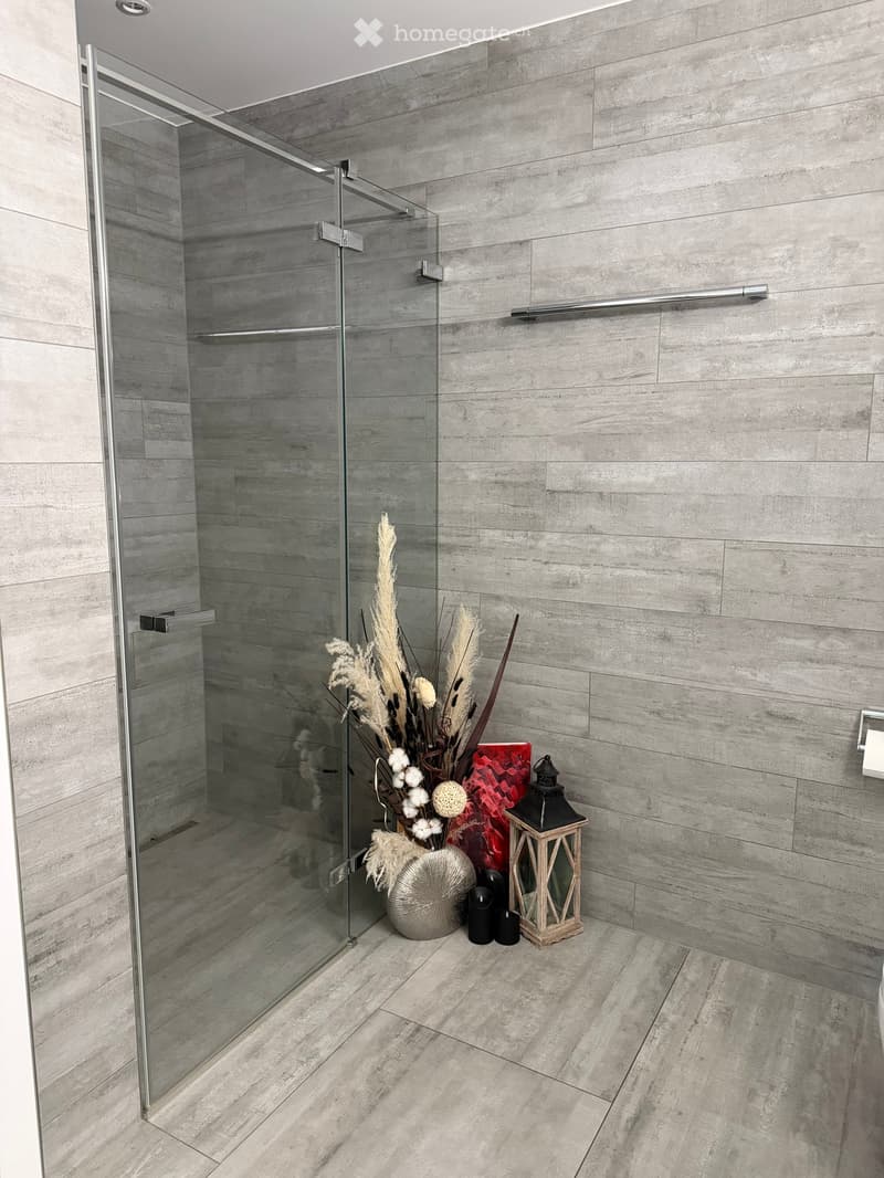 Modern bathroom with a glass shower enclosure, grey tiled walls, and a decorative flower arrangement on the floor near the shower.