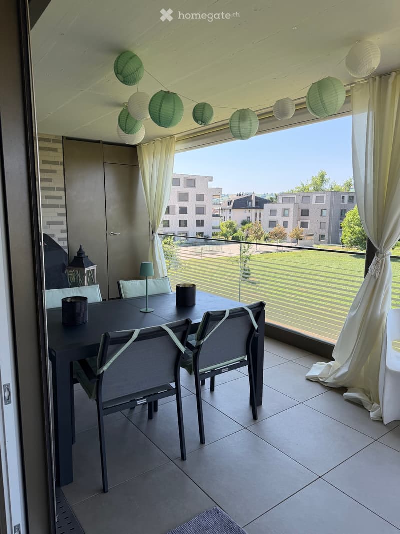 Balcony with a dining table, chairs, a small lamp, and a view of the surrounding buildings and greenery.