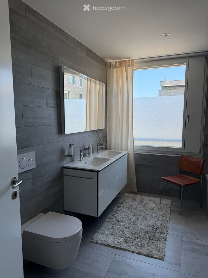 Modern bathroom with double sink, toilet, large mirror, and a small chair near the window.
