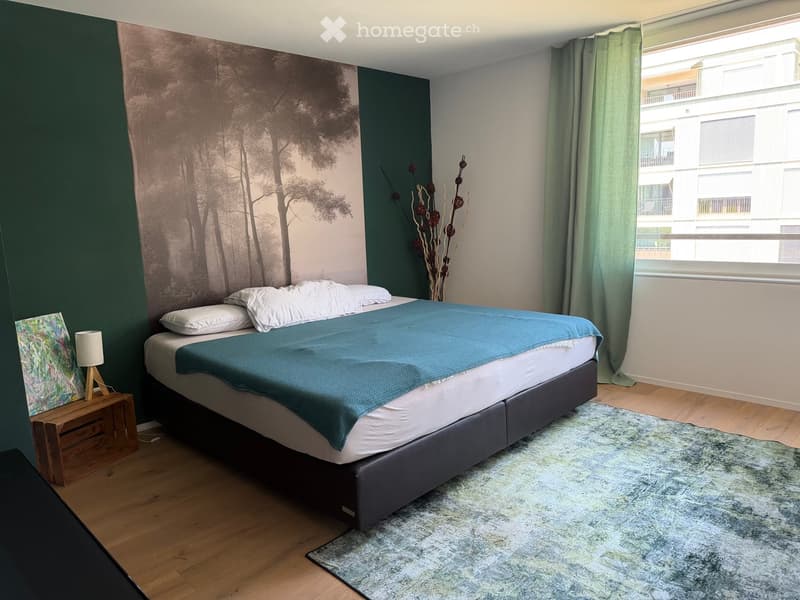 Modern bedroom with a large bed, nightstand, lamp, painting, and decorative vase. The room features wooden flooring and large windows with green curtains.
