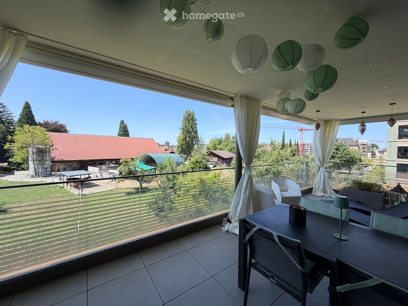 Balcony with a view of a park, featuring a table, chairs, a lamp, and hanging paper lanterns.