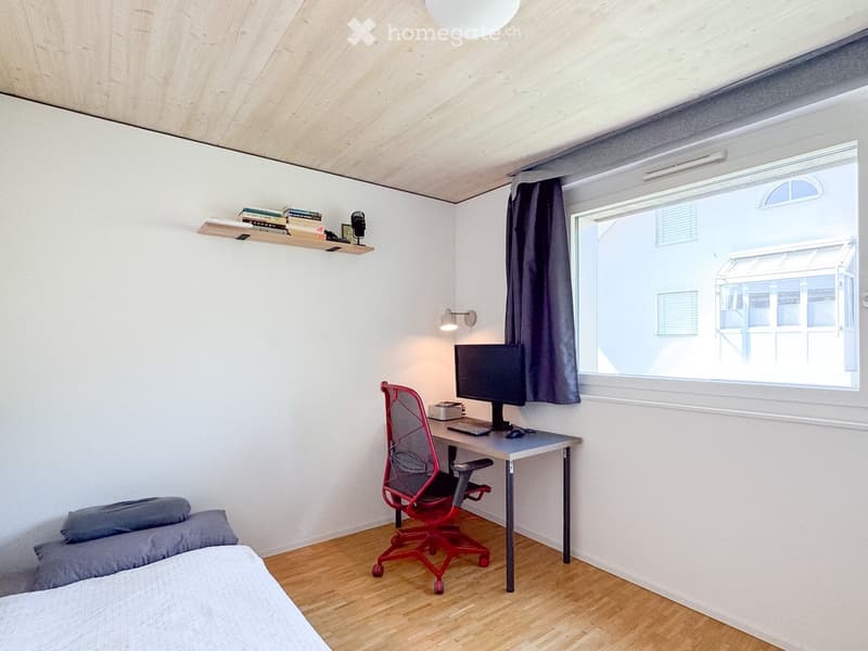 A modern, compact bedroom with a bed, desk, and chair. The room features a large window with a dark curtain, a floating shelf with books, and a sleek, red office chair.