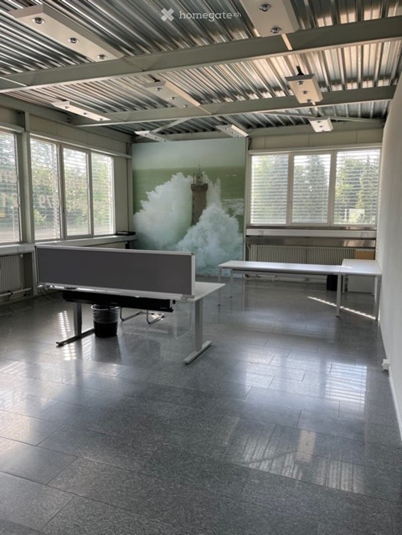 Empty room with large windows, industrial-style ceiling, and a few basic furniture pieces including a desk and a bench.