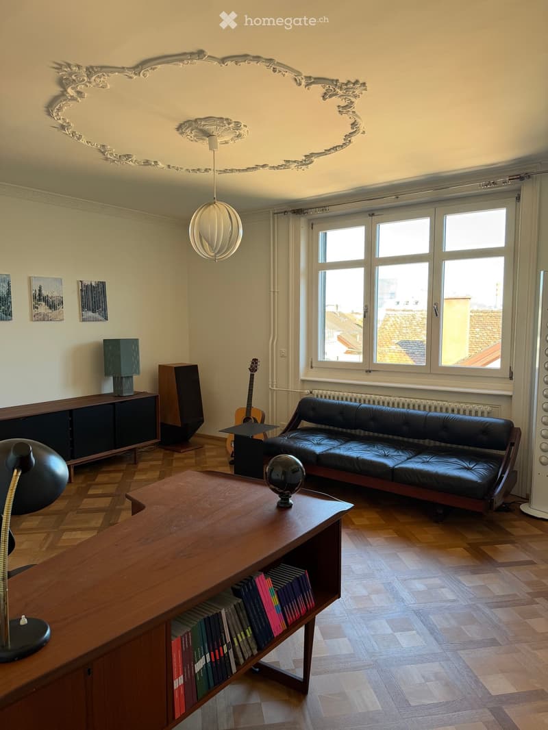 Living room with wooden flooring, large windows, a black leather sofa, a wooden coffee table with a globe, a bookshelf with books, a guitar, a lamp, and a speaker system.