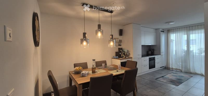 A modern dining and kitchen area with a wooden dining table, six chairs, a bowl of fruit, a bottle, and a cup on the table. The kitchen features white cabinets, an oven, a fridge, and various appliances.