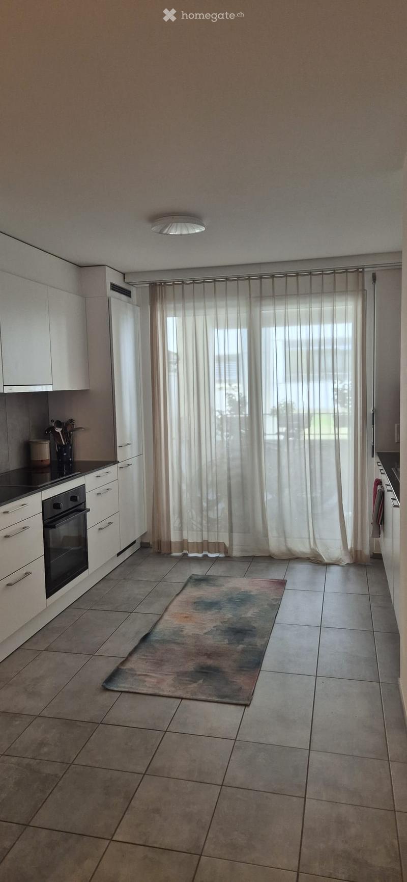 Modern kitchen with white cabinets, an oven, refrigerator, and a large window with sheer curtains. A colorful abstract rug is placed on the tiled floor.