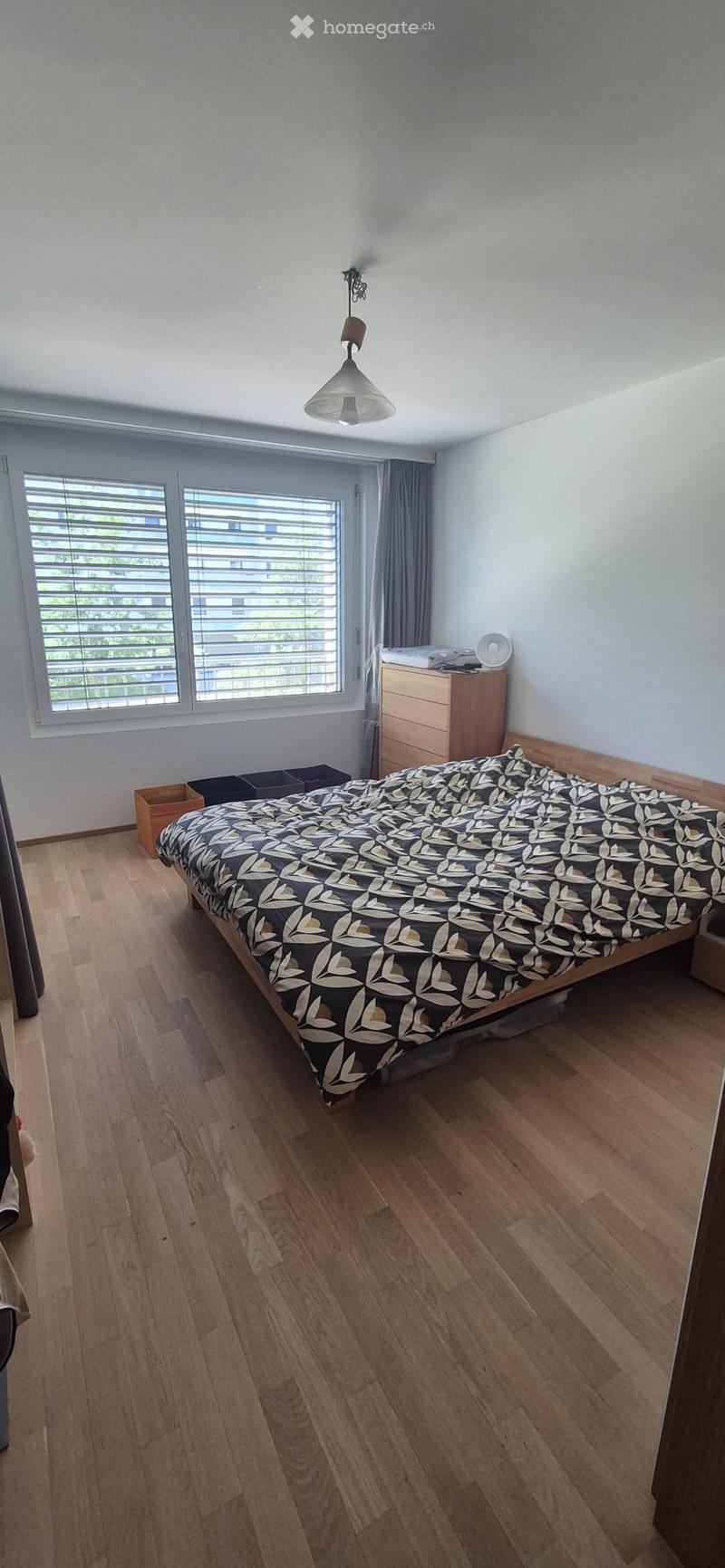 Bedroom with wooden flooring, large window with blinds, bed with patterned duvet, nightstand, and storage boxes.