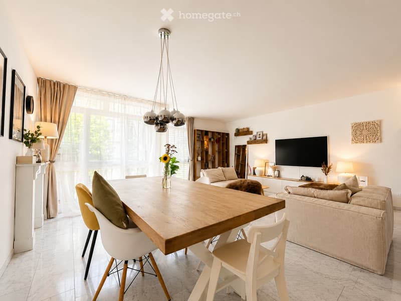 Bright living room with a large wooden dining table, white chairs, a beige sofa, a TV, and decorative items including vases and lamps.