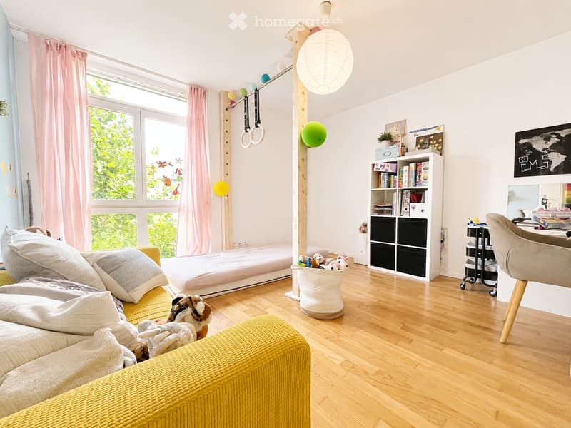 Bright, modern bedroom with wooden flooring, large windows with pink curtains, a low bed, a yellow sofa, a bookshelf, a hanging ball, and a small round table with a chair.