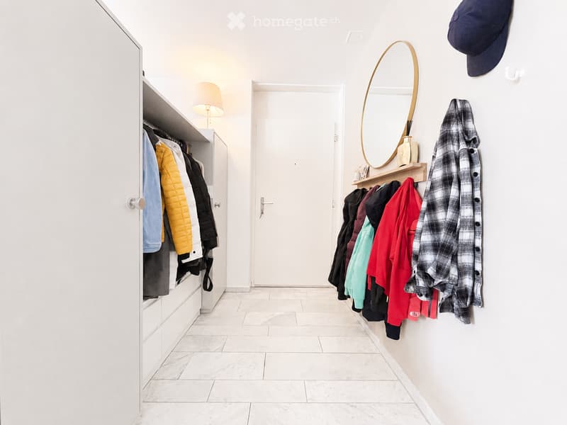 A modern hallway with white walls, marble flooring, built-in closet with hanging clothes, coat rack with jackets, and a round mirror on the wall.