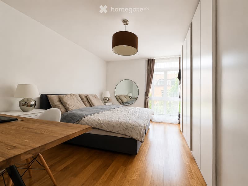 Modern bedroom with a wooden floor, a large bed with pillows and a quilt, a wooden table with a chair, a round mirror, and several lamps.