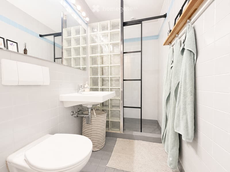 Modern bathroom with white tiles, a toilet, sink, mirror, and a glass-enclosed shower area. Includes a woven basket and a towel on a wooden rack.