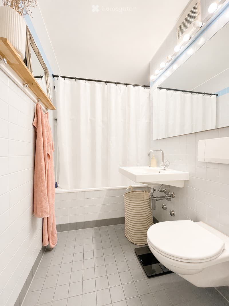 Modern bathroom with white tiles, a sink, toilet, shower curtain, and a towel hanging on the wall. A mirror with built-in lighting is above the sink, and a woven basket is placed on the floor.