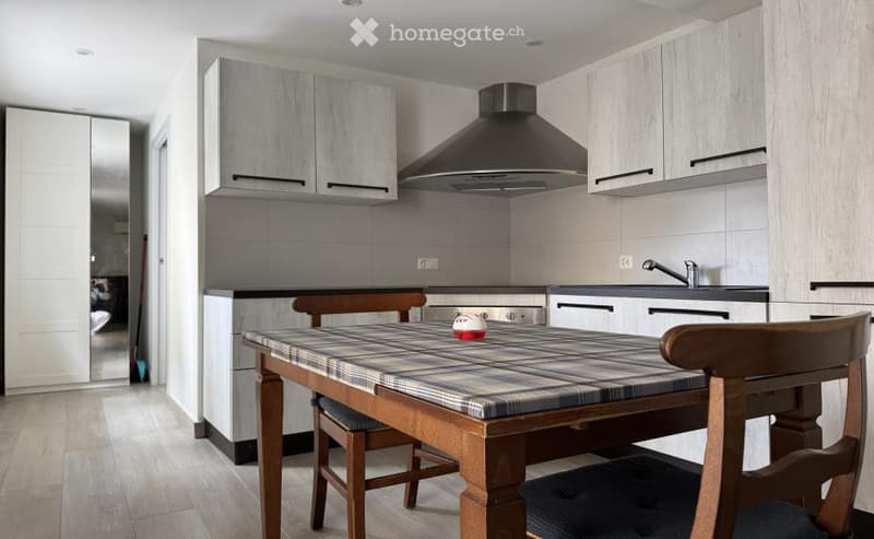Modern kitchen with light grey cabinets, a central dining table with chairs, and a stainless steel range hood above the stove.