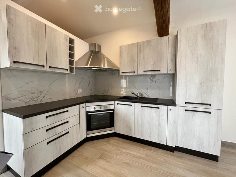 Modern kitchen with light wood cabinets, black countertops, stainless steel oven, sink, and range hood. Marble backsplash and wooden flooring.