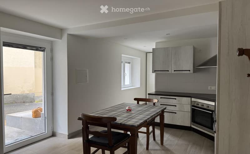 Modern kitchen with grey cabinets, a dining table with chairs, and a large window. The floor is light wood, and the space is well-lit with natural light from the window and sliding glass door.