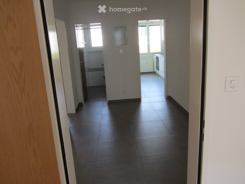 Empty hallway with tiled floor, leading to a kitchen and bathroom. The kitchen has white cabinets and a sink, while the bathroom has a shower and tiled walls.