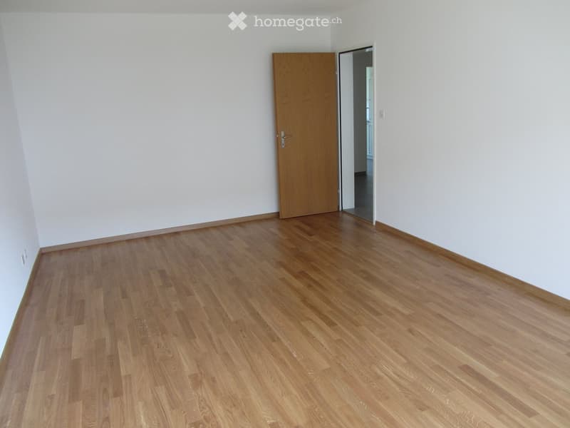 Empty room with light wooden flooring and white walls, featuring a closed wooden door and an open doorway leading to another space.