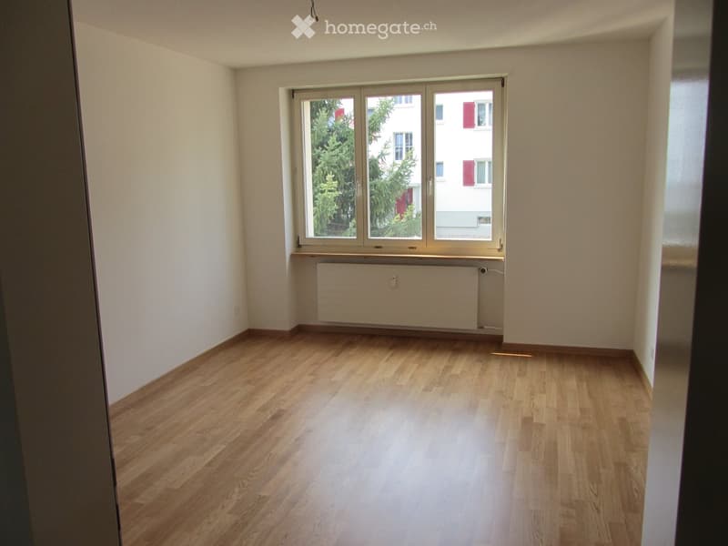 Empty room with wooden flooring, white walls, and a large window with a radiator below it.