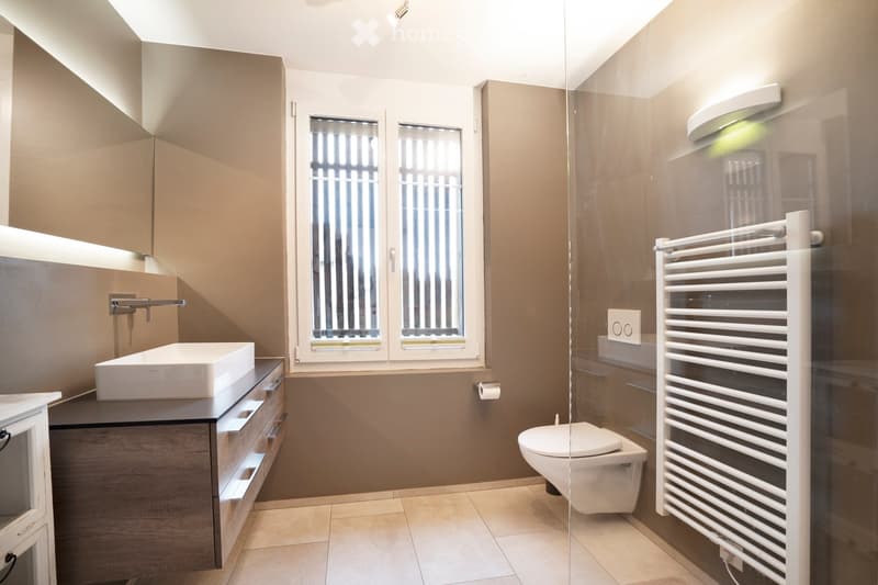 Modern bathroom with a white sink, toilet, radiator, and mirrored cabinet. Features large windows with blinds and warm lighting.