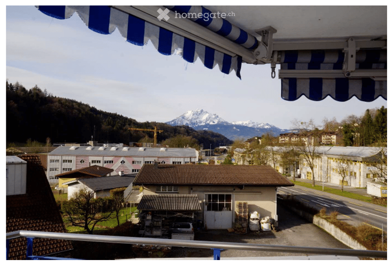 Balcony with a view of a town, industrial buildings, and mountains in the background. The balcony has a blue and white striped awning.