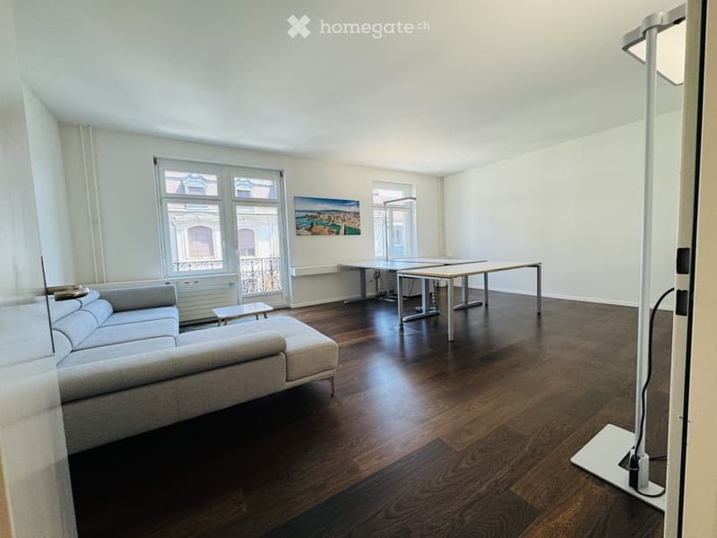 Bright living room with wooden flooring, large windows, a grey sectional sofa, a coffee table, and a dining table with chairs.