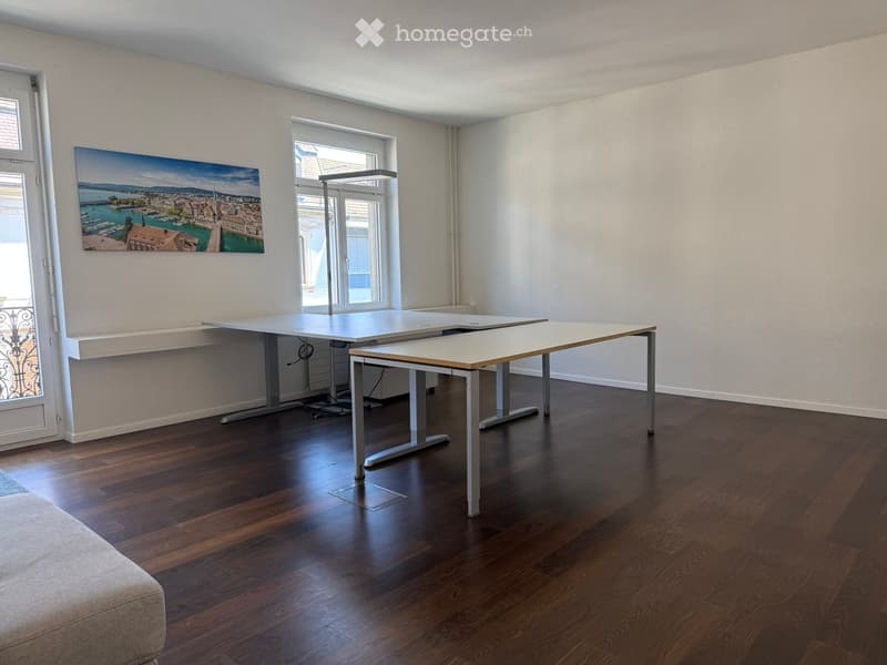 Bright, unfurnished room with wooden flooring, large windows, and a modern table setup. Features a large painting on the wall and a sleek lamp near the window.