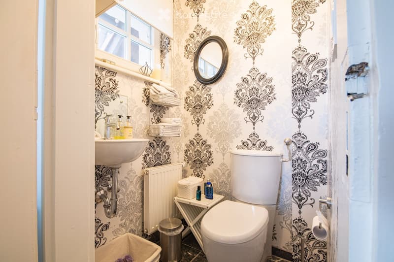 Elegant bathroom with ornate patterned wallpaper, a white sink, toilet, mirror, radiator, and a small stool holding toiletries.