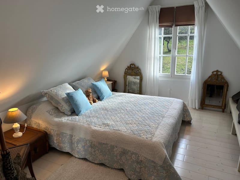 Cozy attic bedroom with wooden floors, large windows, a neatly made bed with pillows and a quilt, two nightstands with lamps, a decorative mirror, and a small wooden stool.
