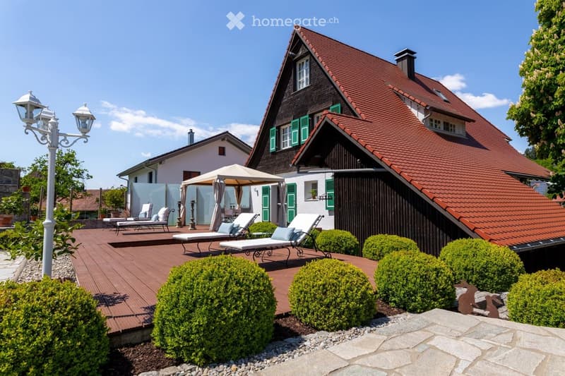 A well-maintained garden area with a wooden deck, lounge chairs, a canopy, and neatly trimmed bushes surrounding the house.