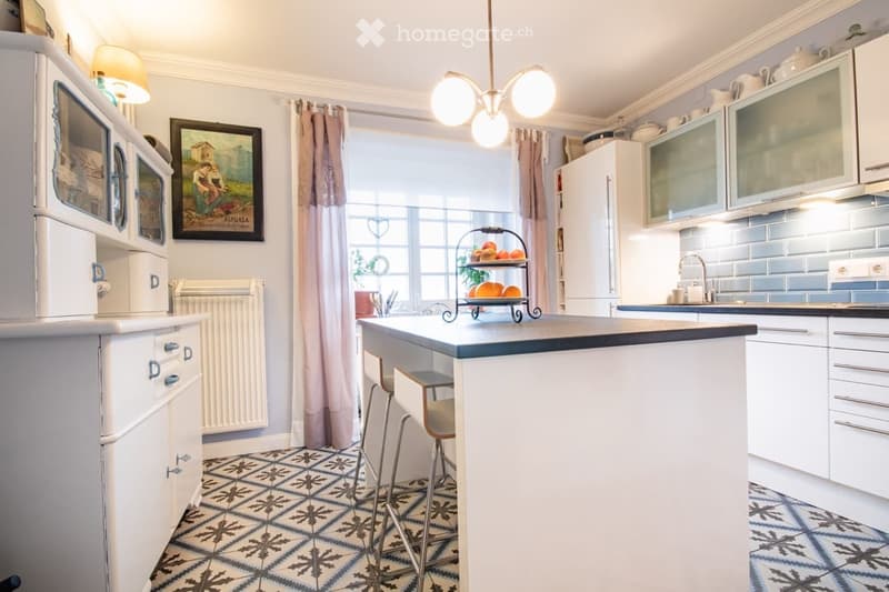 Bright kitchen with white cabinets, a central island, and a vintage-style refrigerator. The space features a patterned tile floor and a window with curtains, complemented by a fruit basket on the island.