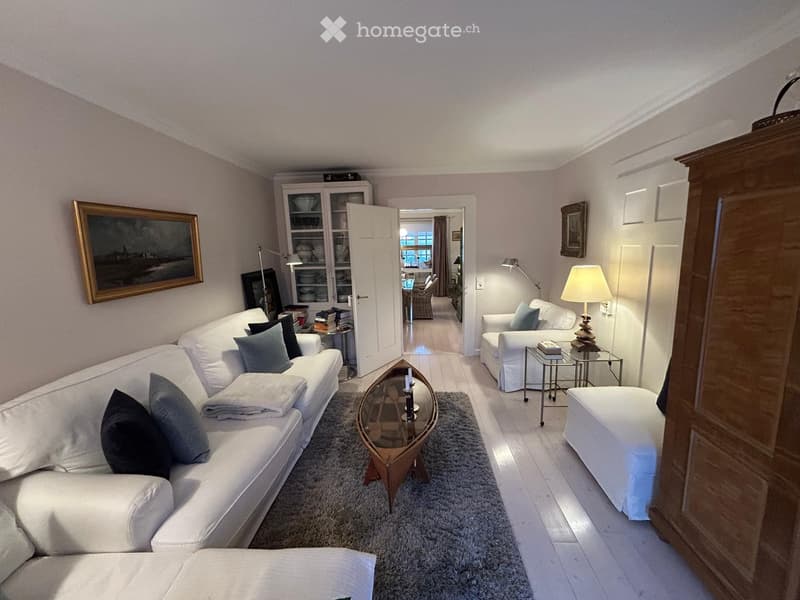 Bright living room with white sofa, coffee table, side tables, lamps, and a cabinet. The room features a grey rug and a view into an adjacent room with a dining area.