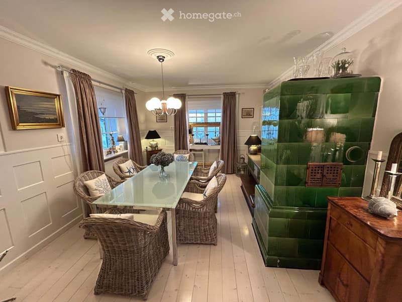 Bright living room with a glass dining table surrounded by wicker chairs, a green fireplace, large windows with curtains, and decorative items including a picture frame and table lamps.
