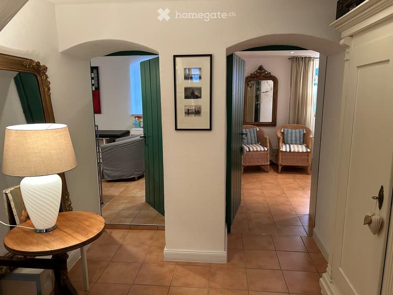 A well-lit hallway with tiled flooring, arched doorways leading to two rooms, and decorative elements including a framed picture and a mirror.