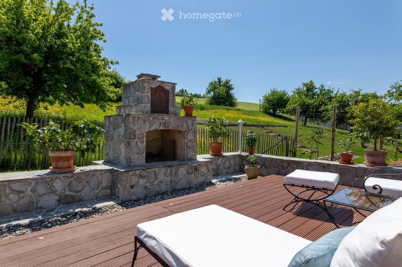 A serene outdoor patio with a stone fireplace, wooden decking, lounge chairs, a small table, and potted plants, surrounded by a lush green landscape.