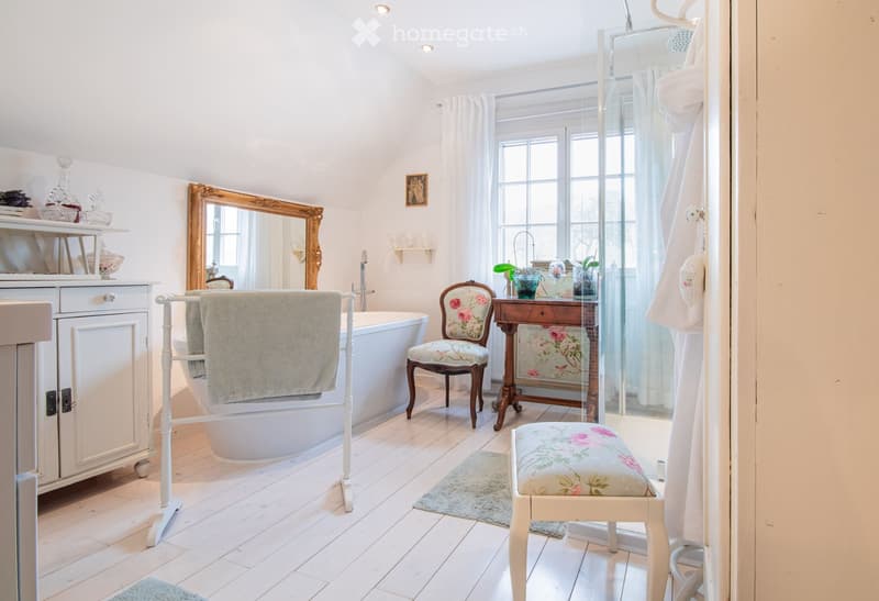 Elegant bathroom with a freestanding bathtub, floral-patterned chairs, a wooden vanity table, a large ornate mirror, and a glass-enclosed shower area.