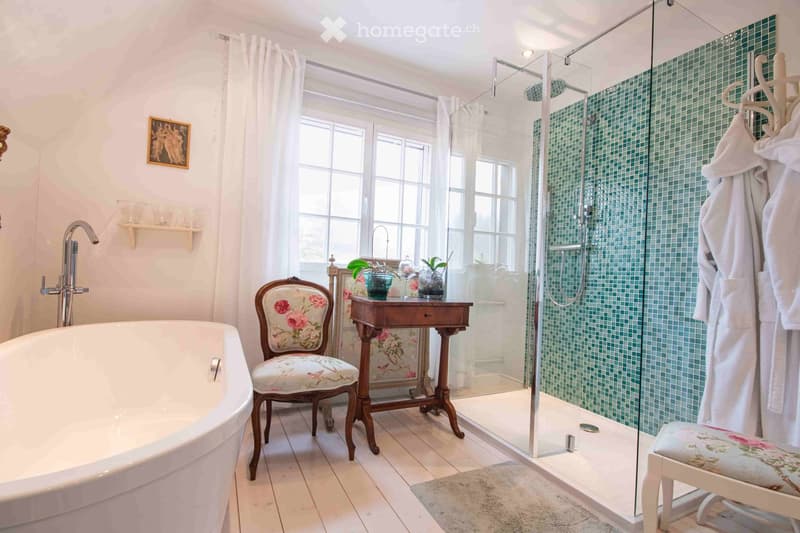 Elegant bathroom with a freestanding bathtub, floral-patterned chair, small wooden table with plants, glass-enclosed shower, and white tiled floor.